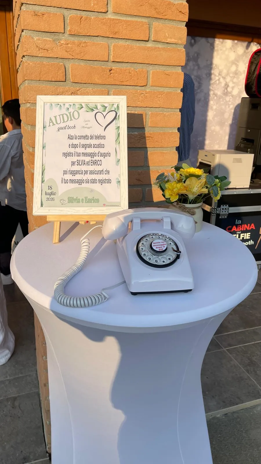 La postazione dell'audio guest book all'ingresso di un ristorante: tavolo alto bianco con telefono vintage, cartello su cavalletto di legno con i nomi degli sposi e un vasetto di fiori gialli