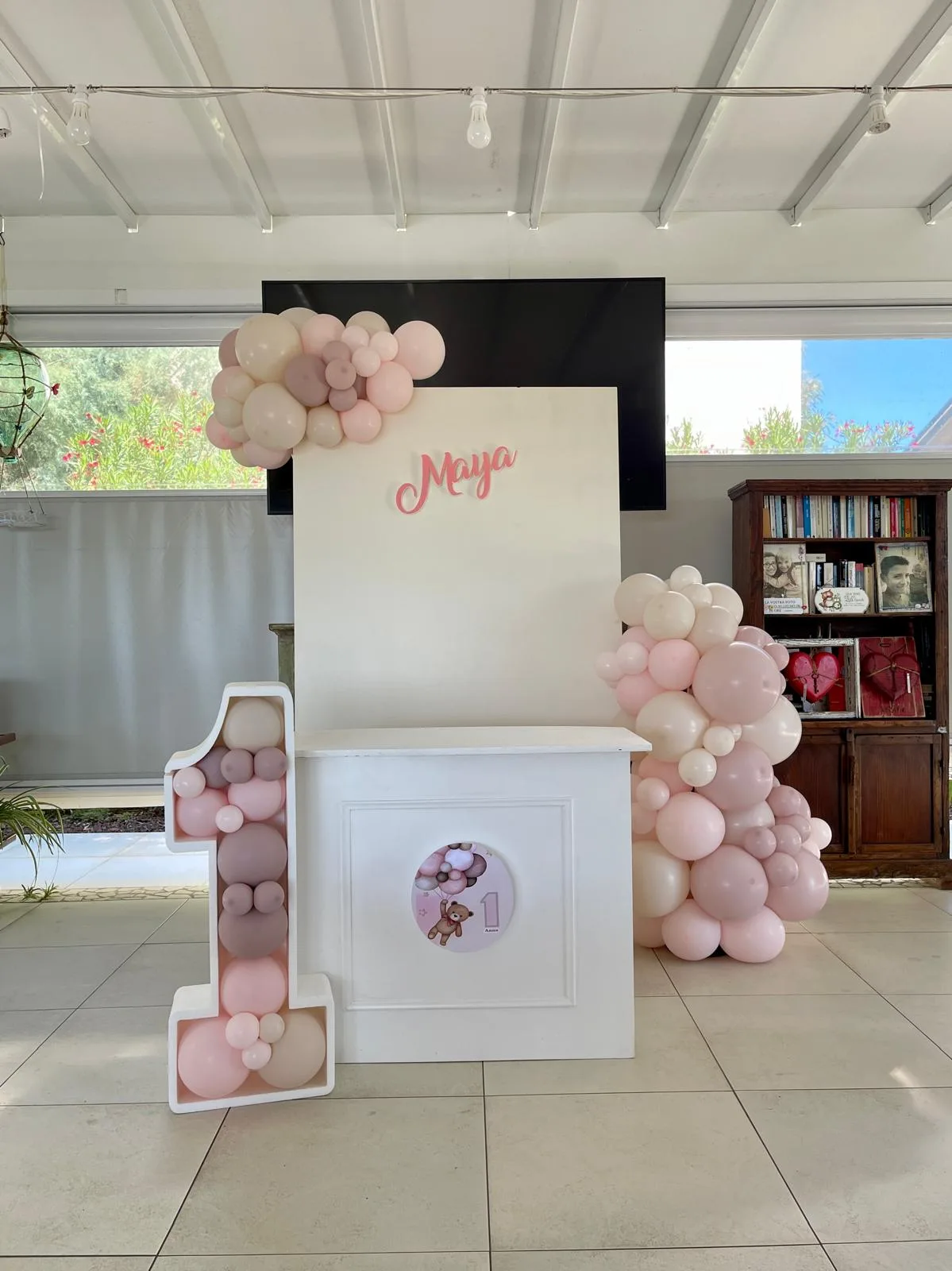 Allestimento per il primo compleanno di Maya: pannello bianco con il nome, numero 1 luminoso riempito di palloncini rosa e tortora e ghirlande organiche ai lati