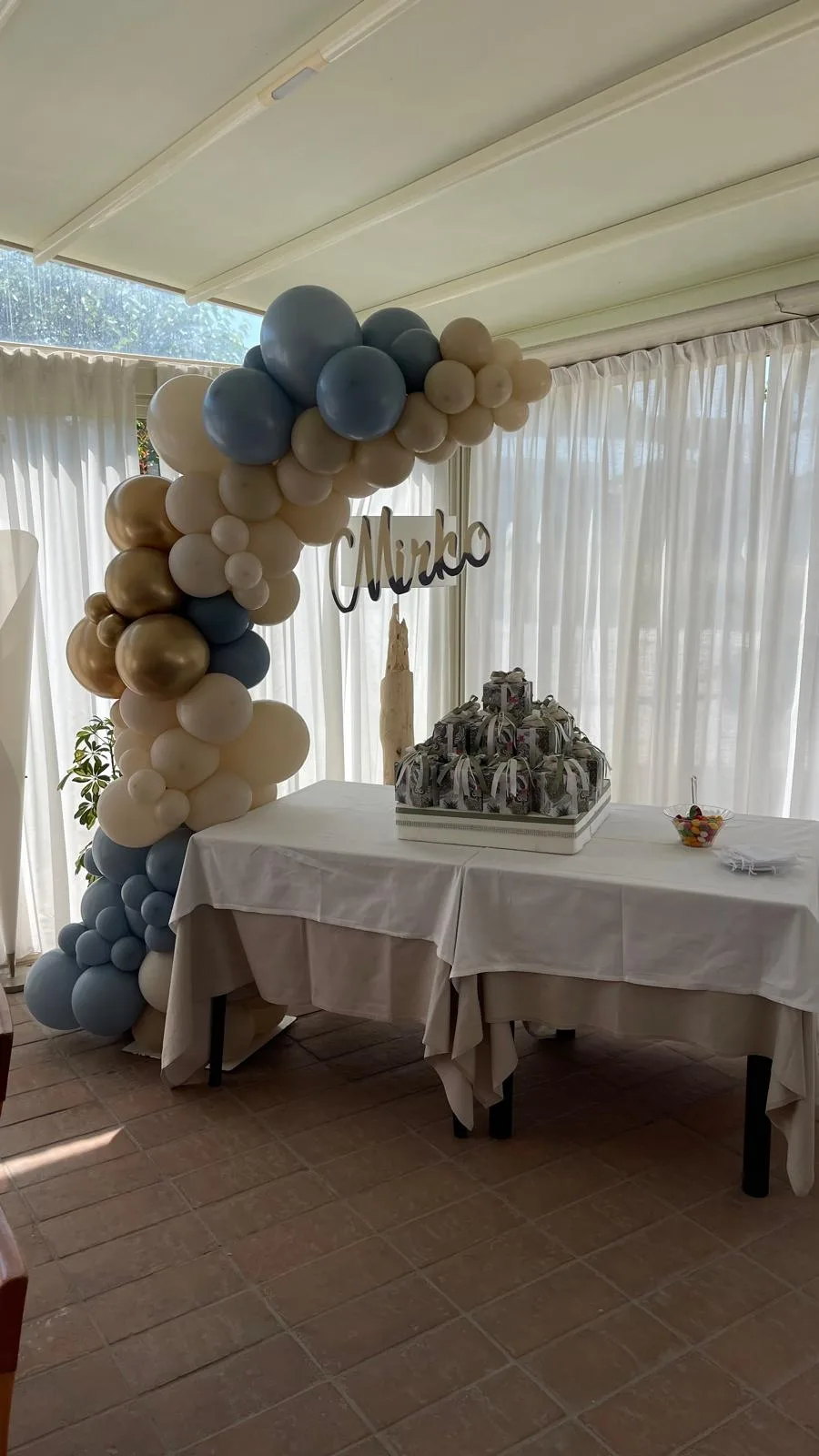 Tavolo dei confetti con arco di palloncini blu, panna e oro, scritta sagomata col nome Mirko e vassoio dei confetti