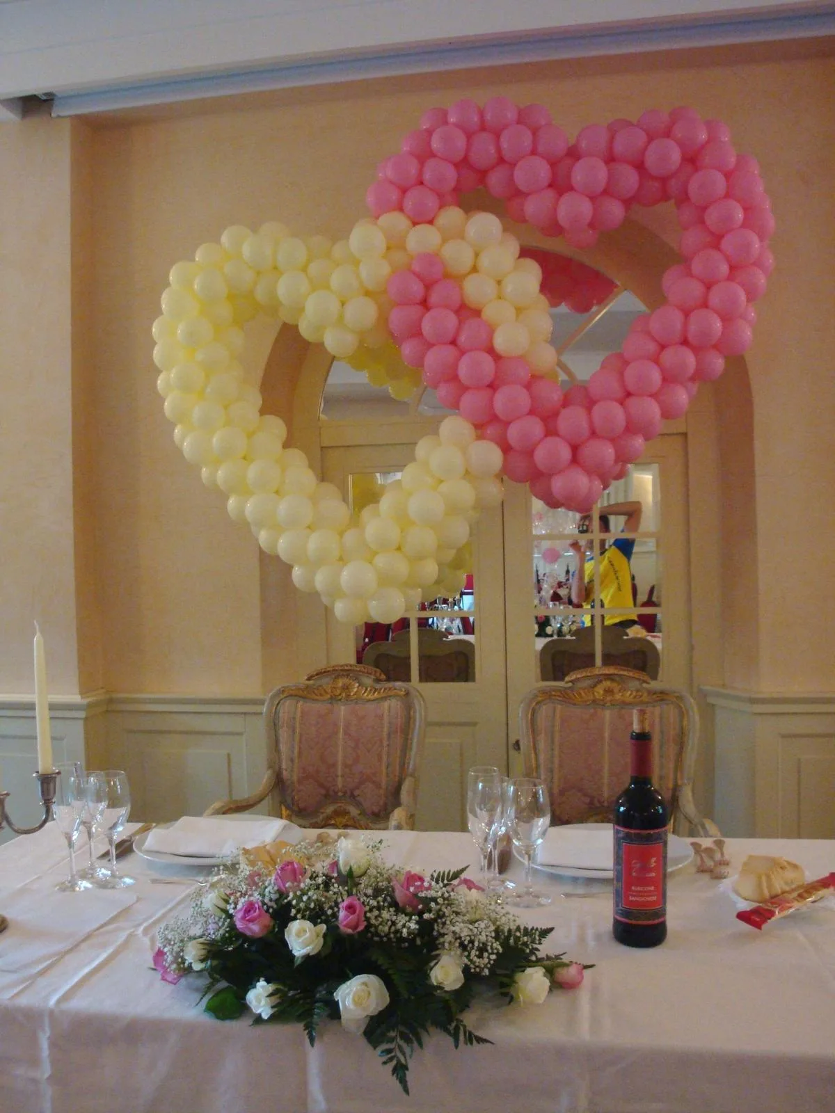 Due cuori di palloncini intrecciati, uno rosa e uno panna, montati alle spalle del tavolo degli sposi apparecchiato con rose e candele