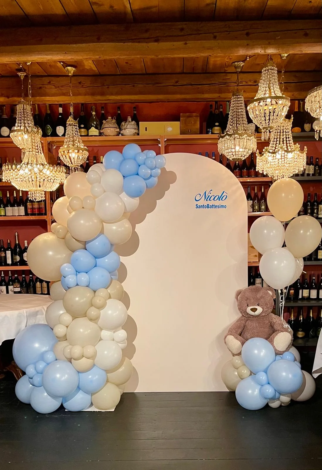Pannello ad arco con la scritta Nicolò Santo Battesimo, ghirlanda di palloncini azzurri e panna, orsetto di peluche e mazzo a elio, allestito in enoteca