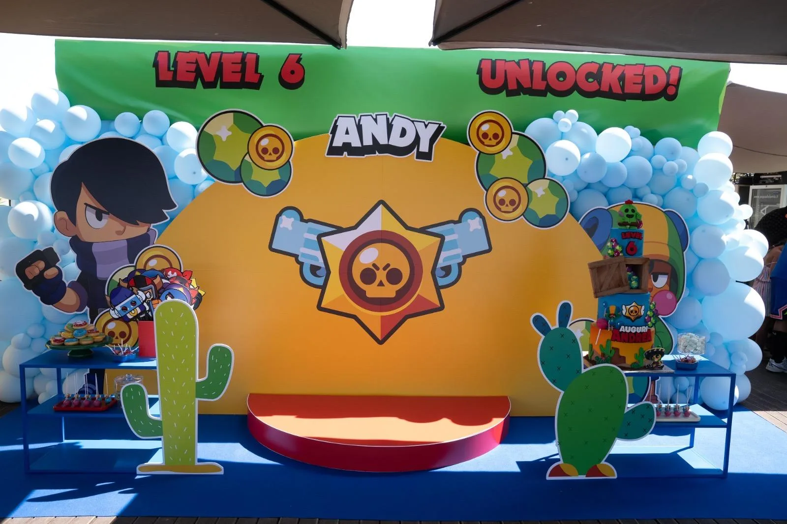 Postazione a tema Brawl Stars con fondale giallo, sagome dei personaggi, ghirlanda di palloncini azzurri e tavoli dei dolci