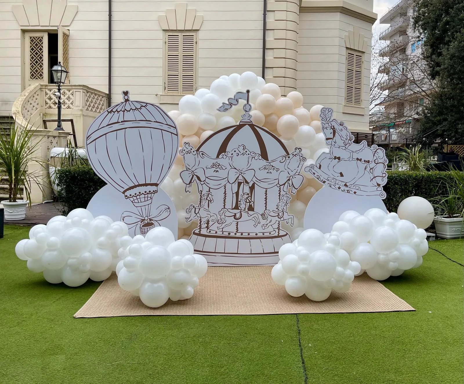 Scenografia in giardino con sagome di giostra e mongolfiera disegnate, nuvole di palloncini bianchi e ghirlanda avorio davanti a una villa