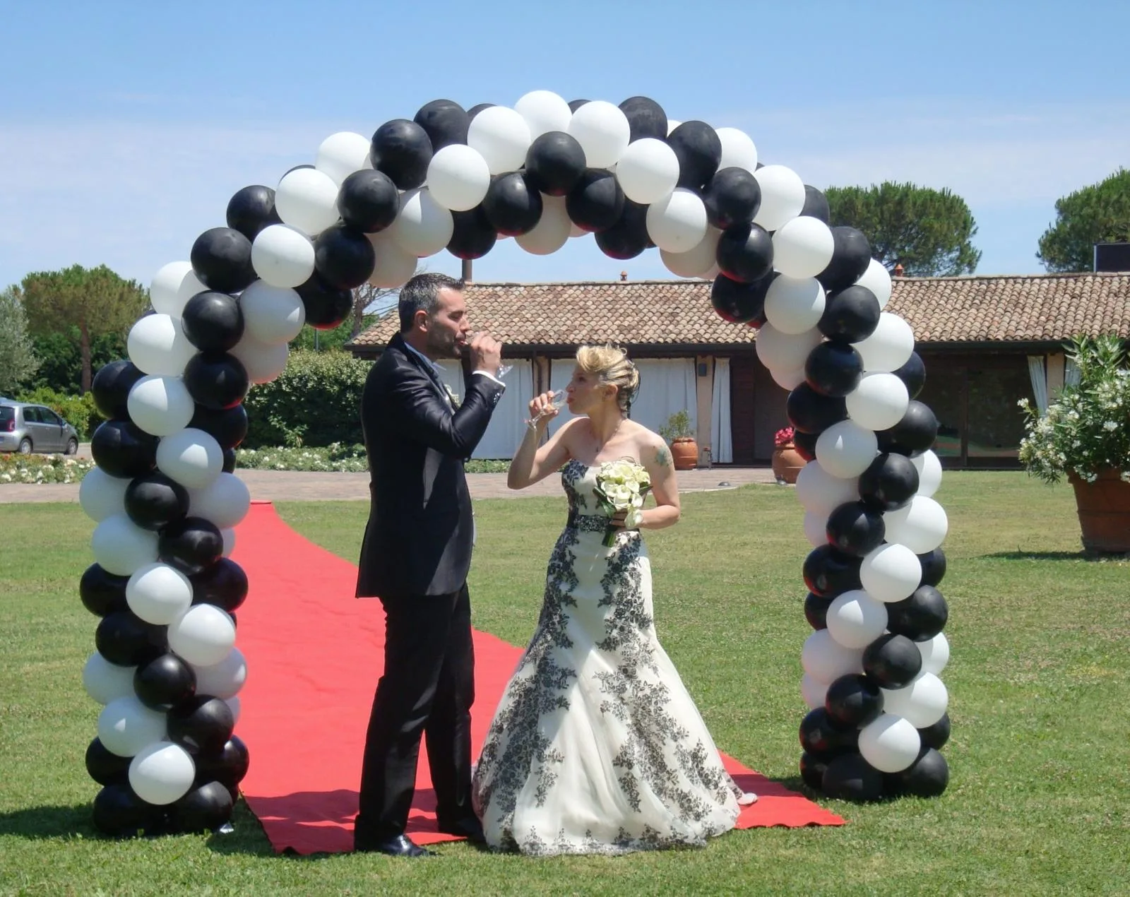 Sposi che brindano sotto un arco di palloncini bianchi e neri montato sul tappeto rosso, in giardino