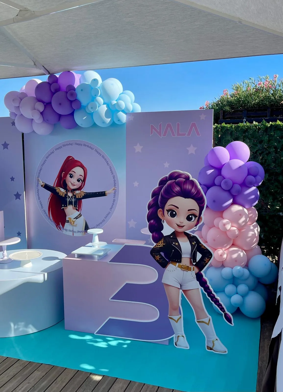 Postazione lilla e azzurra con sagome di una cantante dei cartoni, pannello col nome, banco a gradini e ghirlande di palloncini viola e rosa
