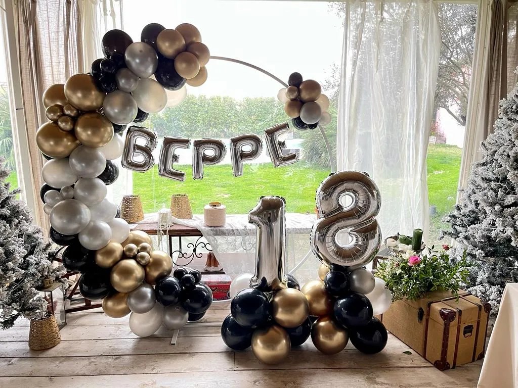Cerchio di palloncini neri, oro e argento con il nome Beppe composto in lettere mylar e numero 18 su colonna, davanti alla vetrata di una sala sul giardino