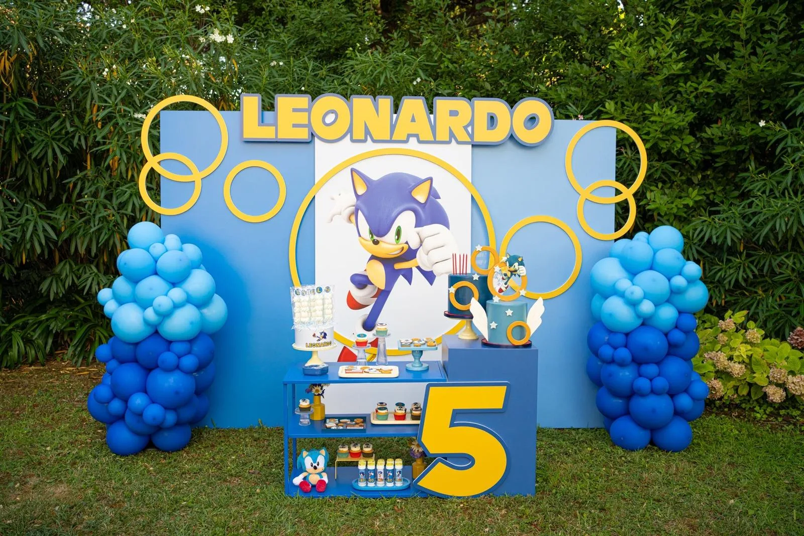 Postazione a tema Sonic in giardino: fondale azzurro con il nome Leonardo in lettere gialle, anelli sagomati, tavolo dei dolci, torte e numero 5