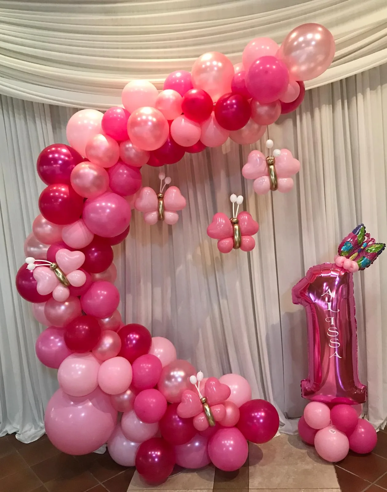 Cerchio di palloncini rosa e fucsia con farfalle e numero 1 in mylar fucsia con il nome Alissa, allestito in sala davanti a un drappeggio bianco