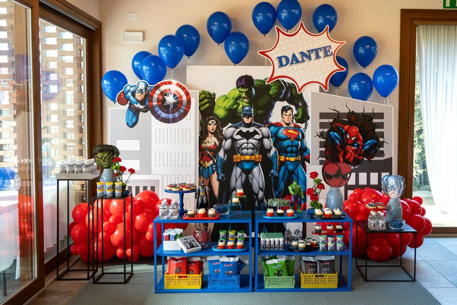 Postazione completa a tema supereroi: fondale con Batman, Superman e Hulk, nuvoletta col nome Dante, tavoli blu dei dolci e colonne di palloncini rossi e blu