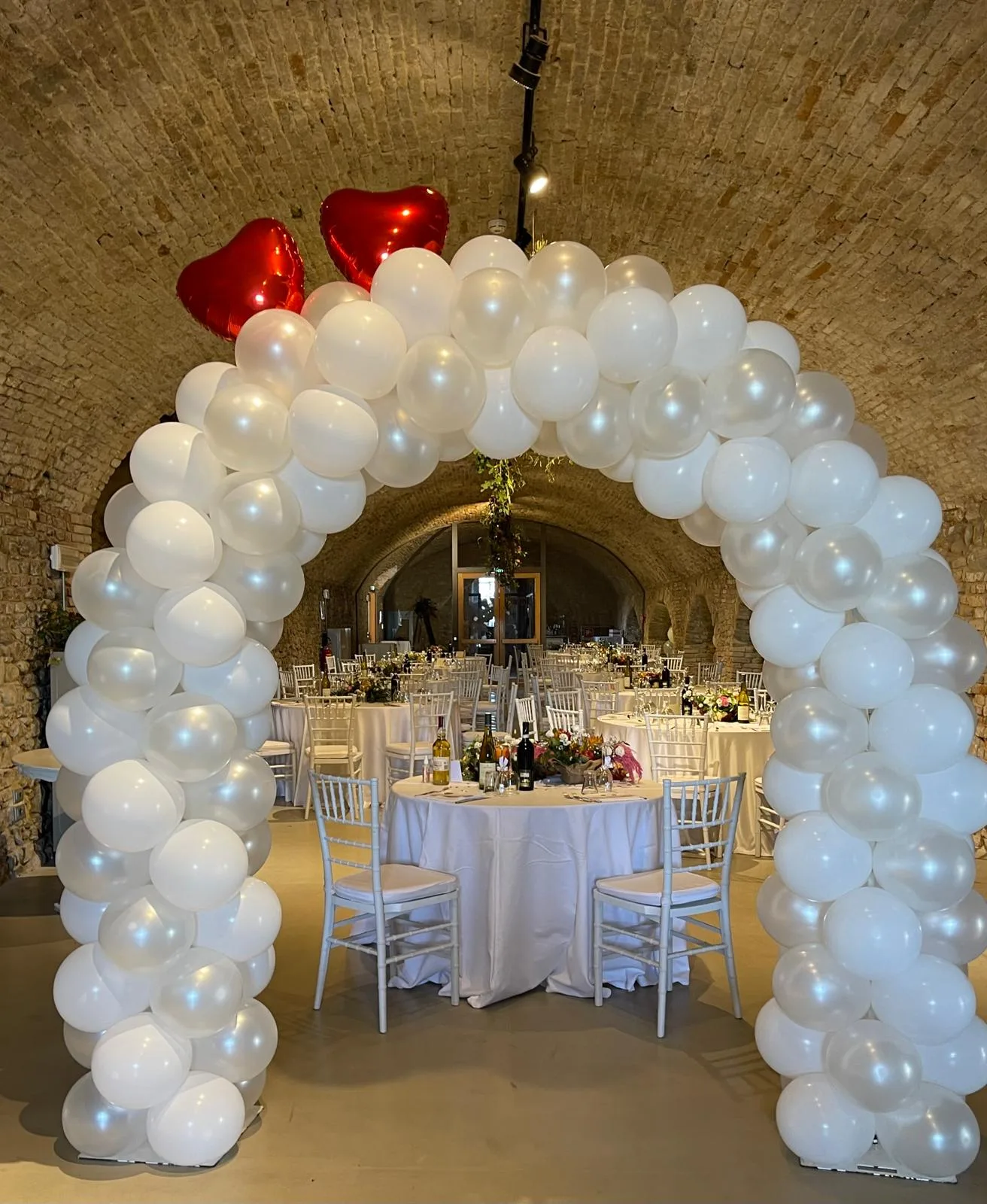 Arco di palloncini bianchi perlati con due cuori rossi in mylar all'ingresso di una sala per ricevimenti con volta in mattoni