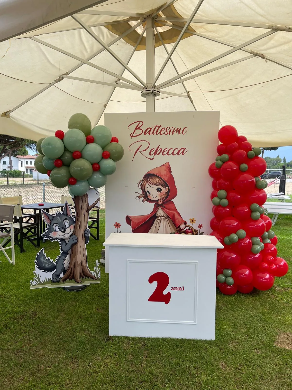Postazione da esterno per un battesimo a tema Cappuccetto Rosso: pannello col nome Rebecca, banco bianco con il numero 2, albero di palloncini e colonna rossa