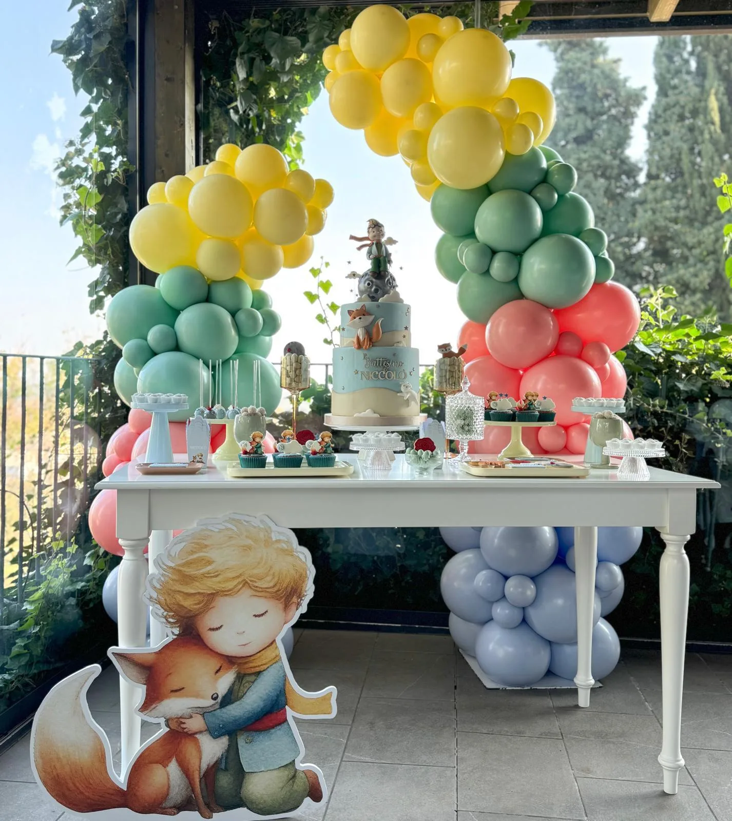 Tavolo dei dolci per un battesimo con doppie colonne di palloncini gialli, verdi e rosa, sagoma del bambino con la volpe e torta a tre piani