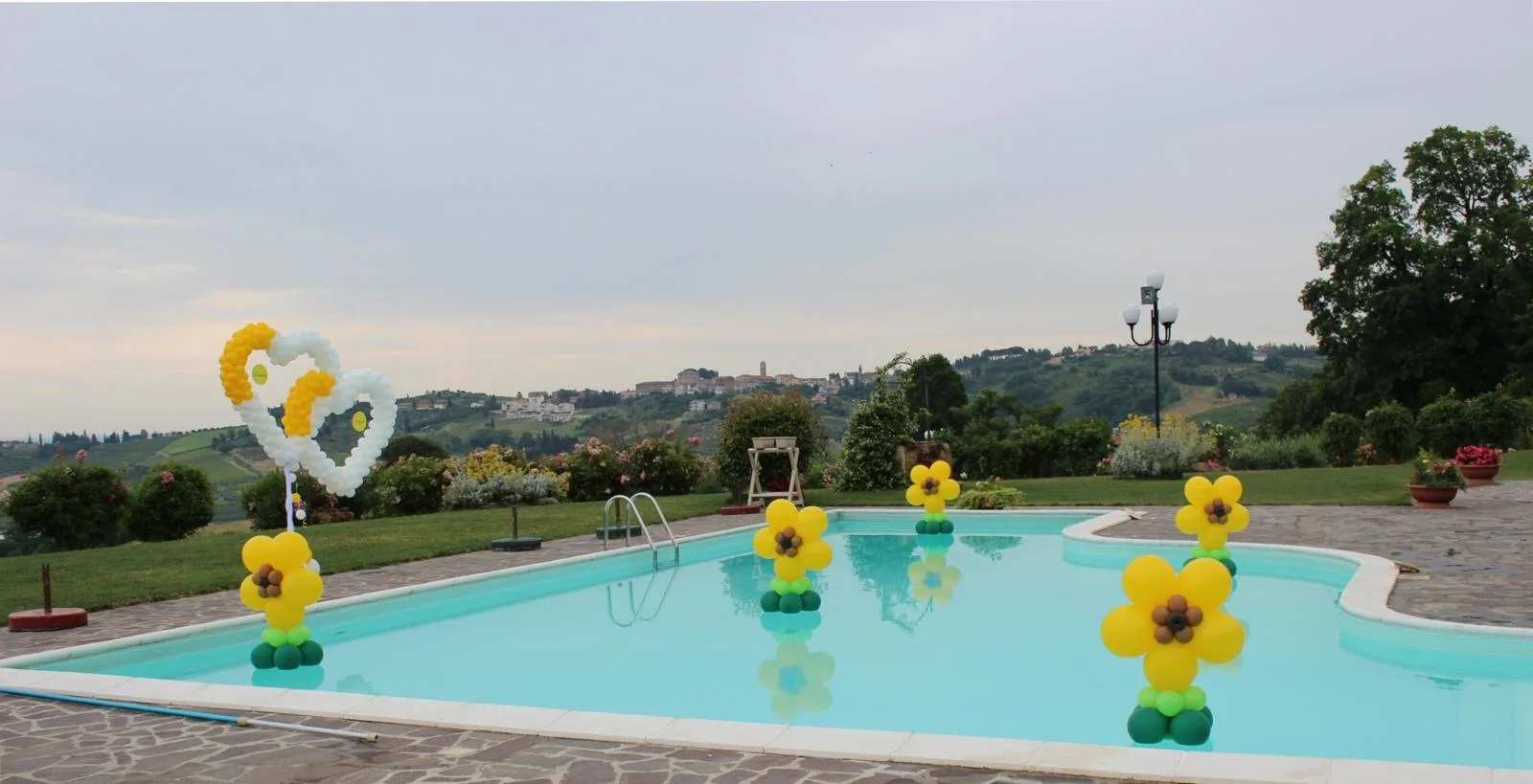 Piscina di un agriturismo sulle colline con fiori di palloncini gialli galleggianti e due cuori intrecciati bianchi e gialli sul bordo