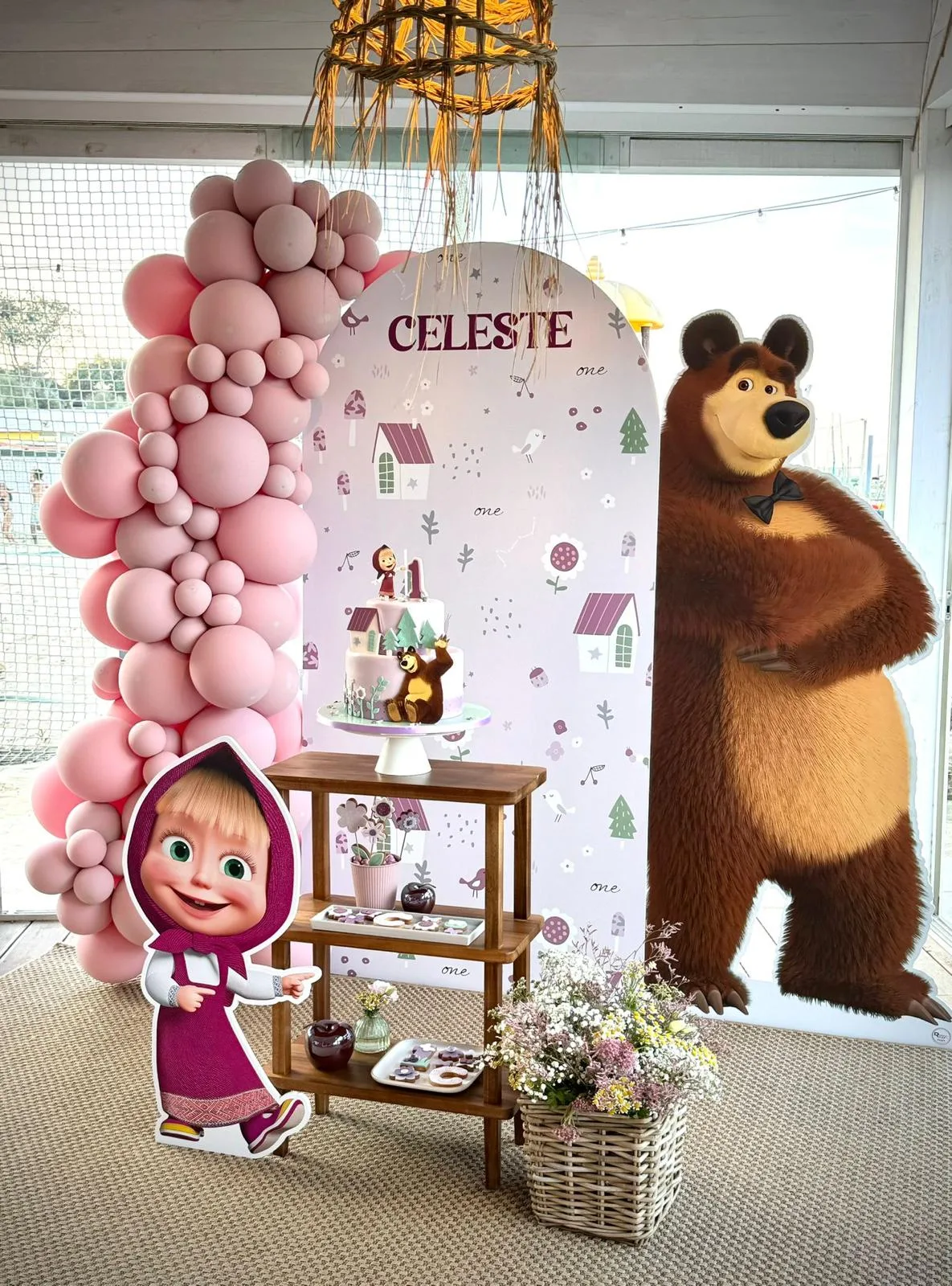 Postazione a tema Masha e Orso: pannello ad arco col nome Celeste, sagome di Masha e dell'orso, ghirlanda rosa, scaletta in legno con torta e dolcetti