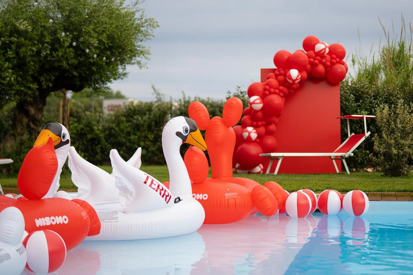 Festa in piscina con fondale rosso e ghirlanda di palloncini rossi e palloni da spiaggia, gonfiabili a forma di cigno e fenicottero in acqua