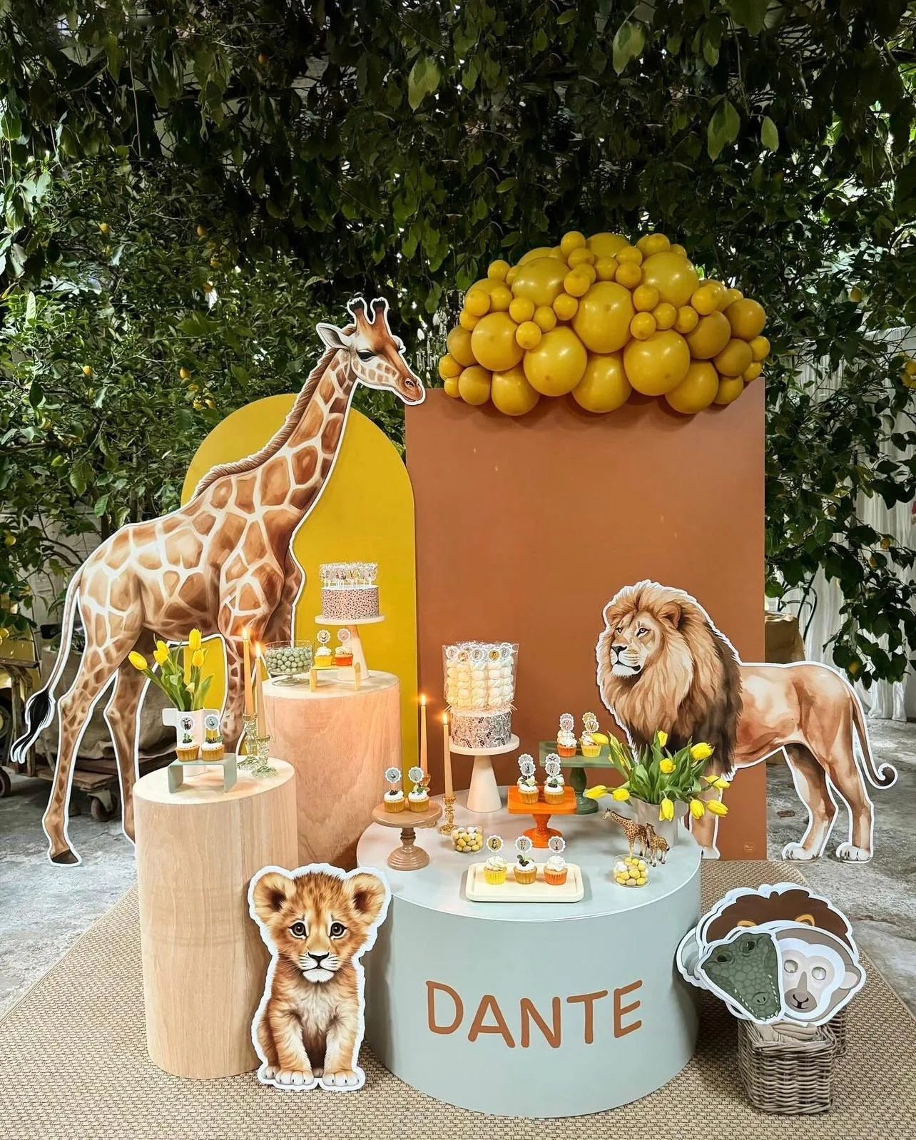 Postazione a tema safari: pannelli ocra e senape, giraffa e leone sagomati a grandezza naturale, nuvola di palloncini gialli e banco tondo col nome Dante