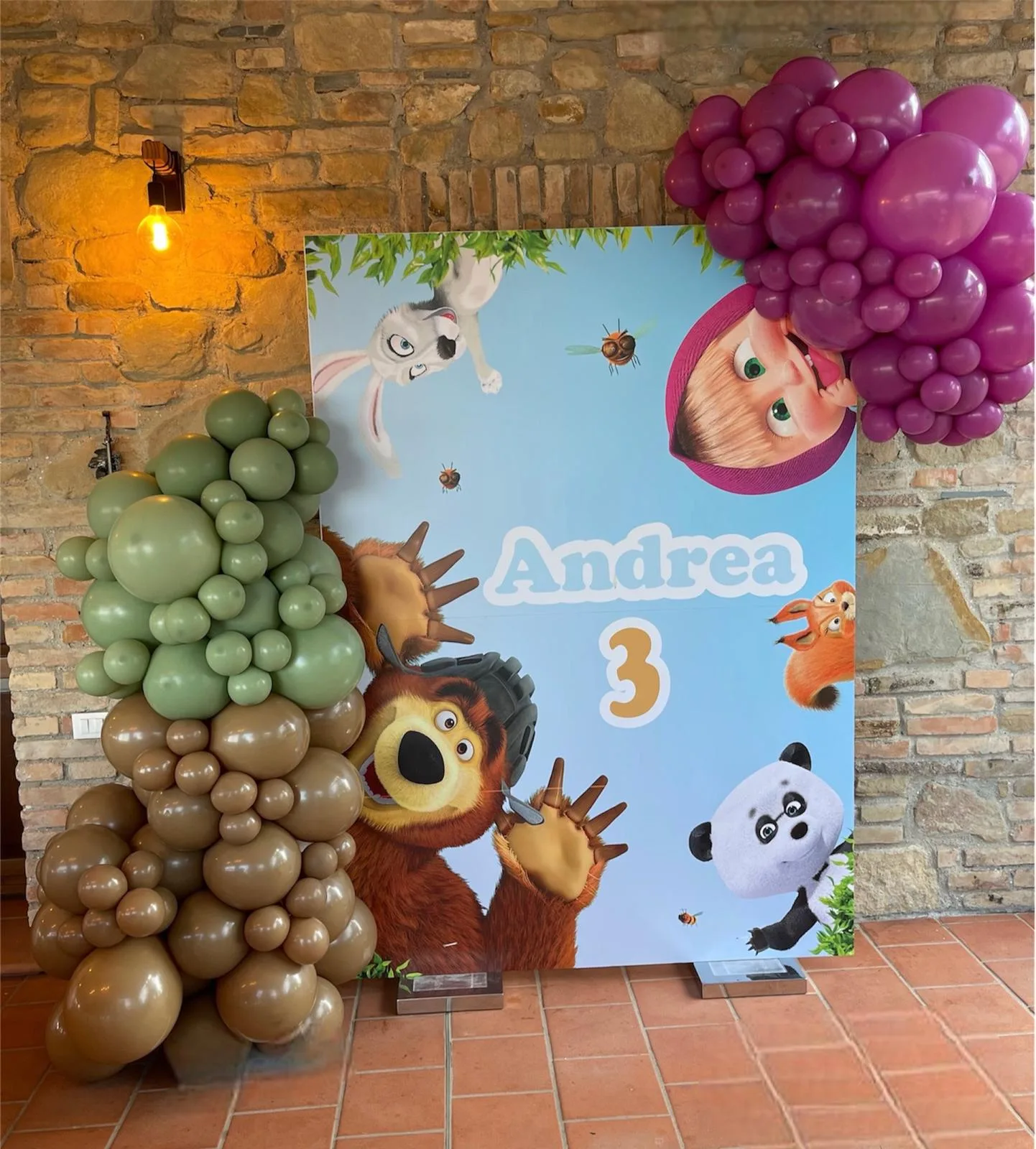 Fondale a tema Masha e Orso col nome Andrea e il numero 3, con colonne di palloncini verde salvia, marrone e fucsia, montato su un muro in sasso