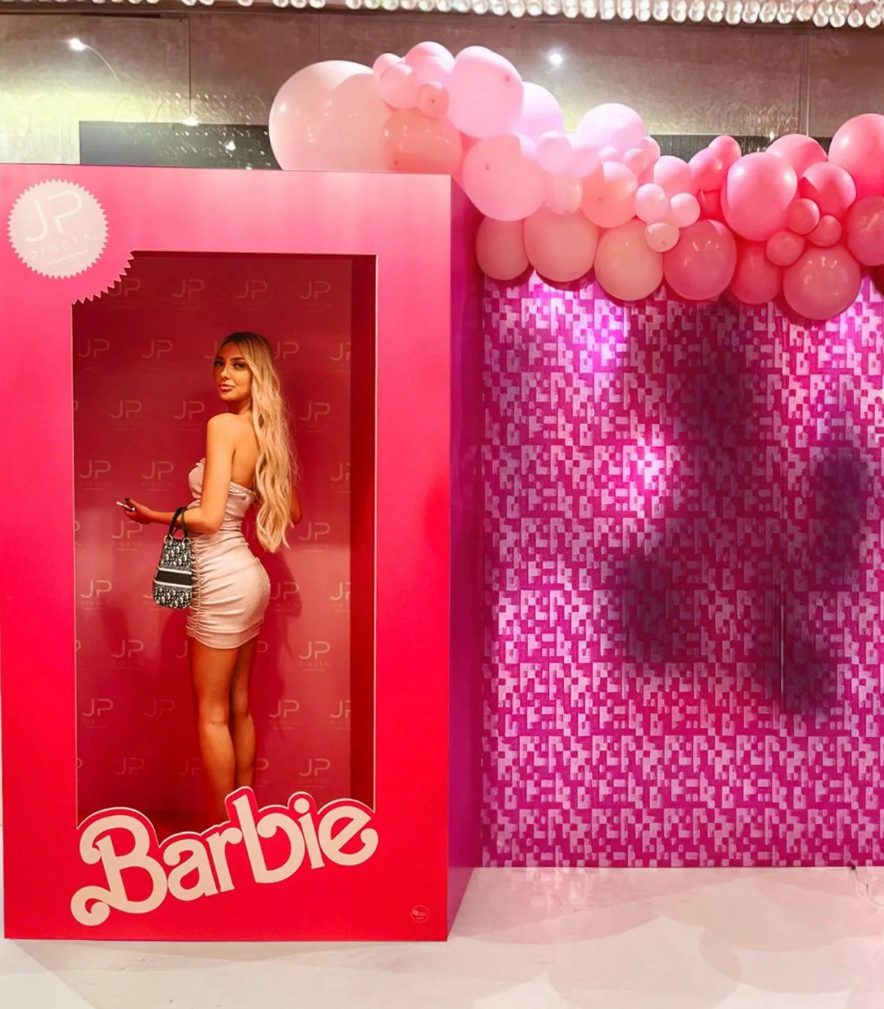 Scatola sagomata a grandezza naturale in stile bambola con una ragazza dentro, accanto a una parete glitter fucsia con ghirlanda di palloncini rosa