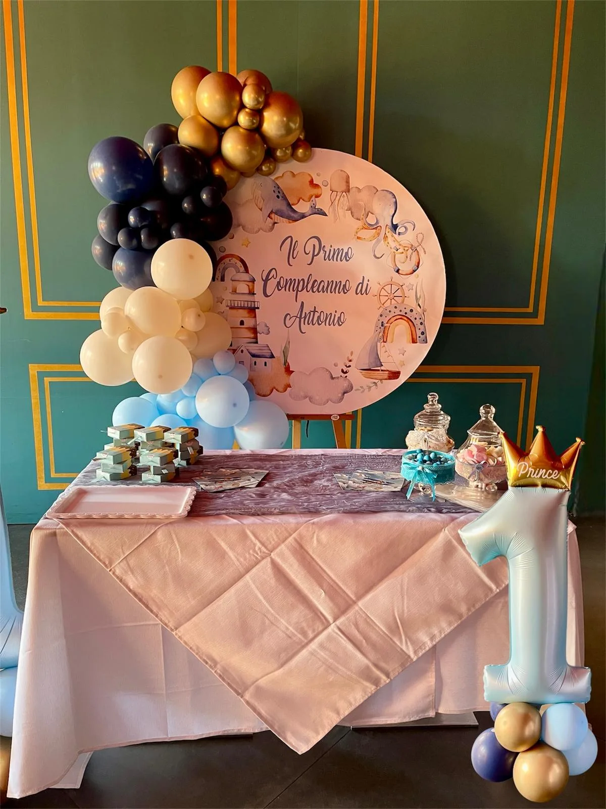 Tavolo dei dolci con pannello tondo stampato Il primo compleanno di Antonio, ghirlanda di palloncini blu, oro e panna e numero 1 celeste