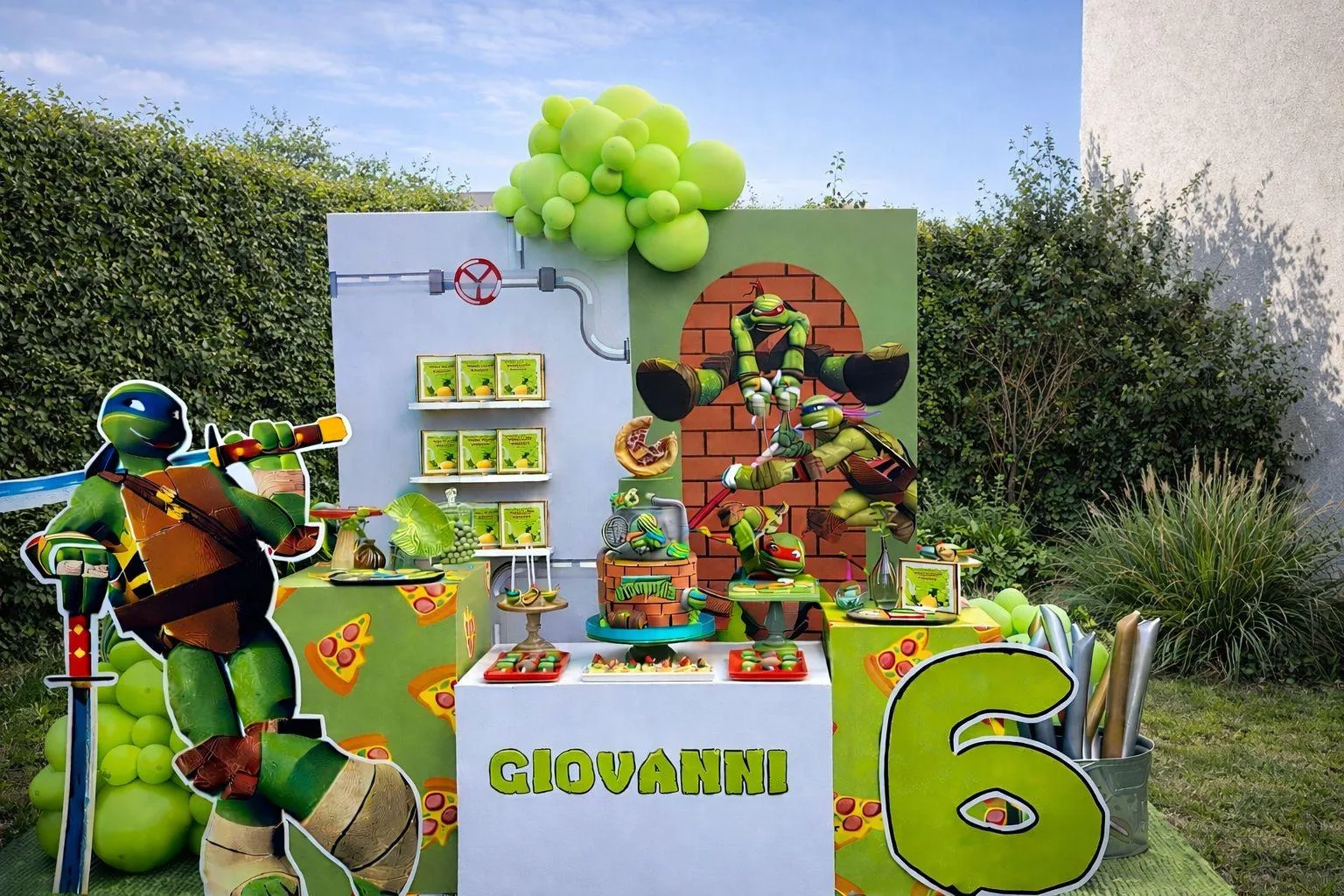 Postazione a tema Tartarughe Ninja in giardino: fondale con muro di mattoni, sagoma di Leonardo, banco bianco col nome Giovanni e numero 6 verde