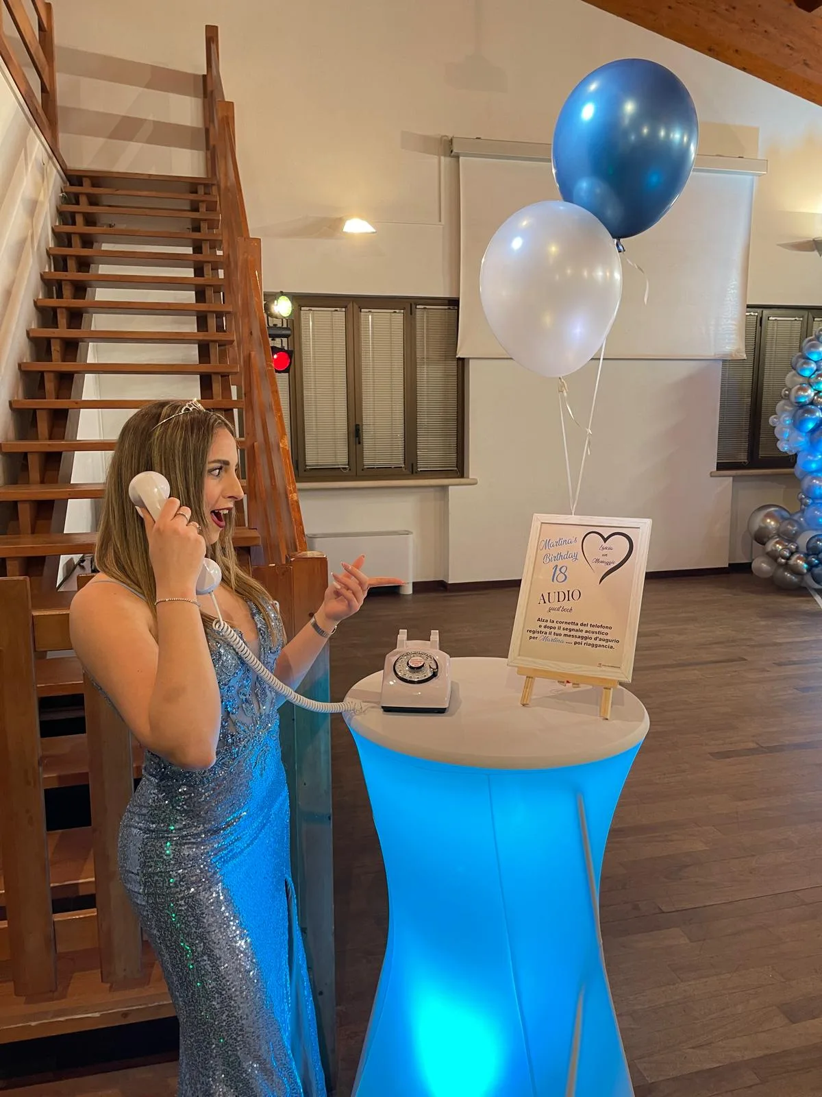 Ragazza in abito da sera con la cornetta all'orecchio che lascia un messaggio all'audio guest book di un diciottesimo, con palloncini azzurri e tavolo illuminato