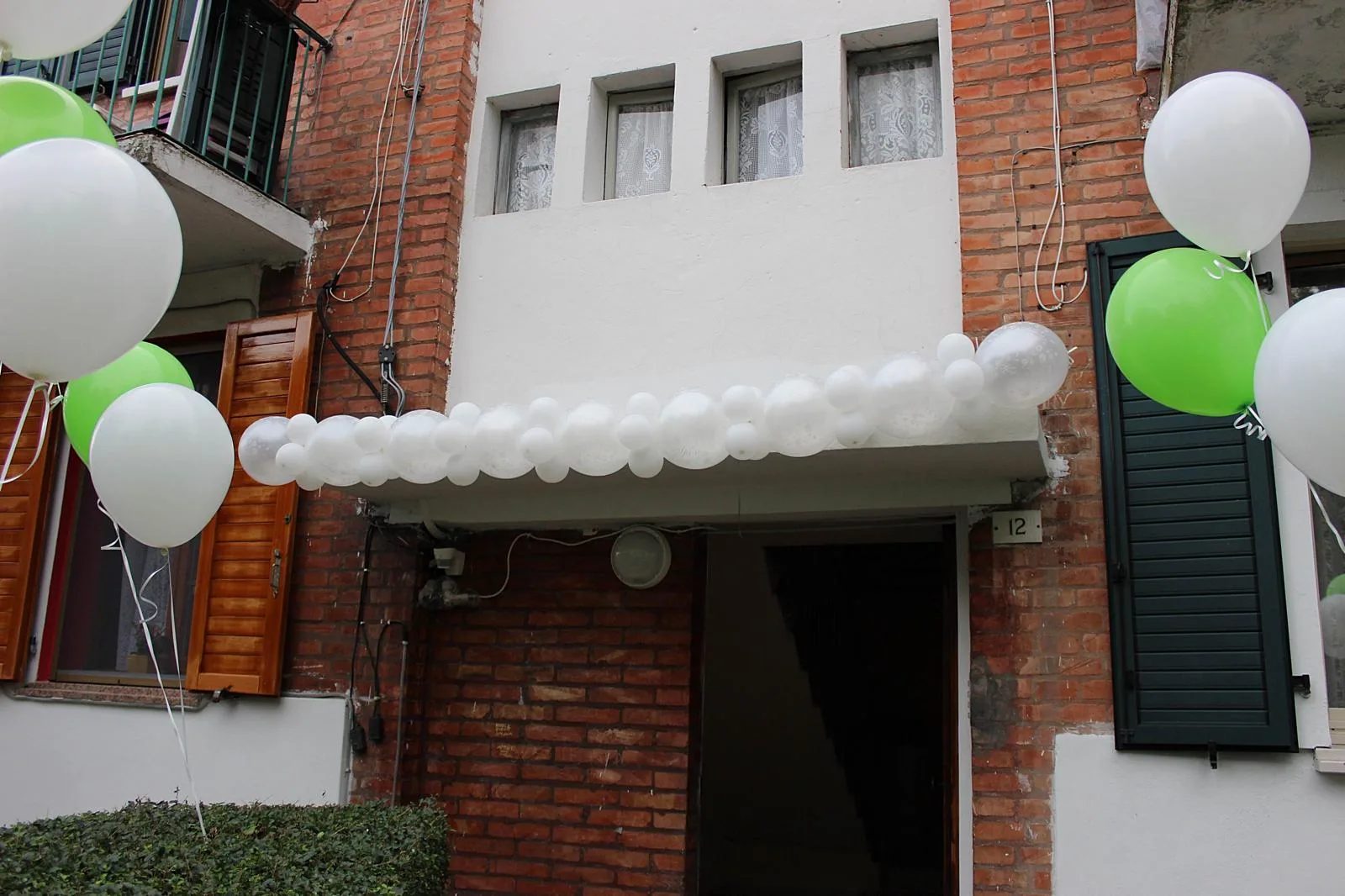 Ghirlanda di palloncini bianchi sopra il portone di una casa, con mazzi di palloncini bianchi e verdi a elio ai lati