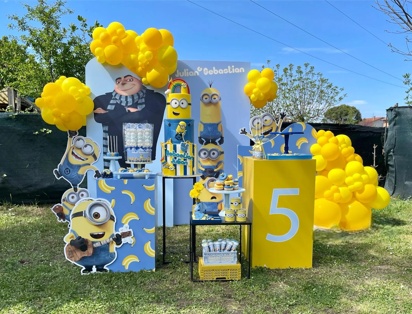 Postazione a tema Minions in giardino: pannello con i nomi Julian e Sebastian, sagome dei Minions, banco con le banane, numero 5 e nuvole di palloncini gialli