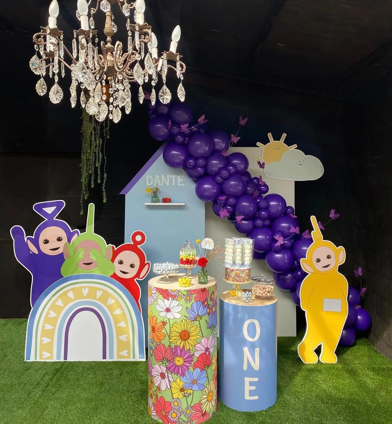 Postazione per il primo compleanno con casetta sagomata col nome Dante, personaggi colorati a grandezza naturale, cilindri con la scritta ONE e ghirlanda di palloncini viola
