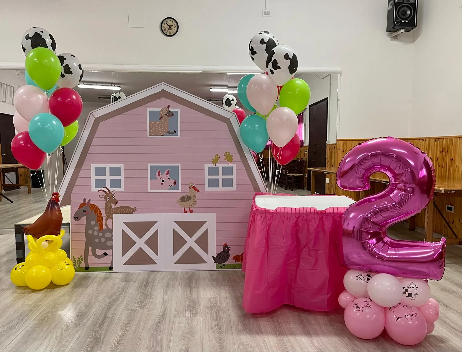 Postazione a tema fattoria in sala: fondale con la stalla rosa e gli animali, mazzi di palloncini mucca, tavolo della torta e numero 2 in mylar fucsia
