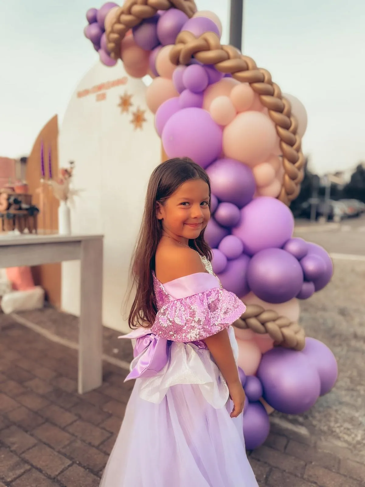 Bambina vestita da principessa davanti all'arco di palloncini lilla, rosa e oro con la treccia intrecciata di palloncini