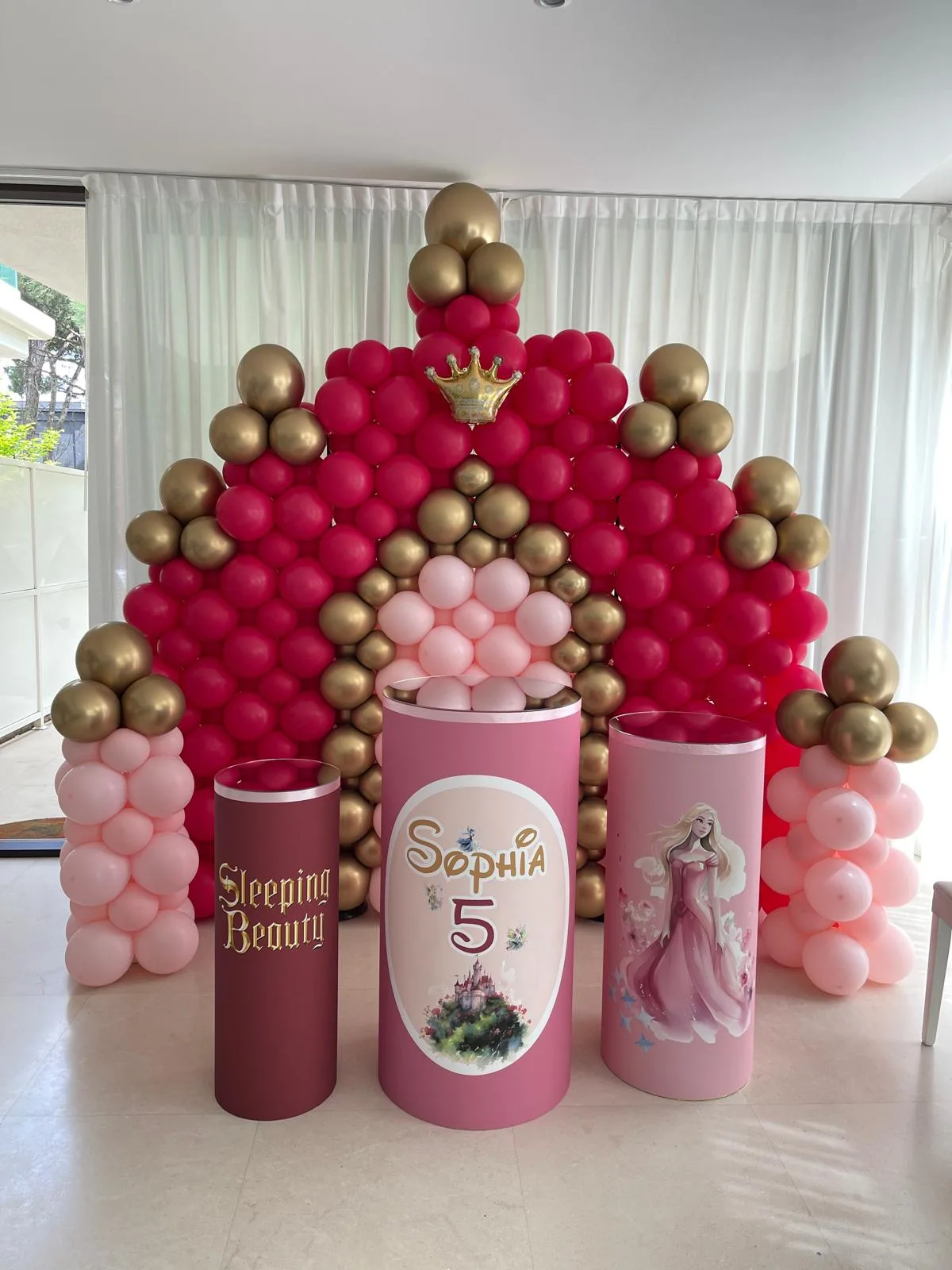 Parete di palloncini a forma di corona in fucsia, rosa e oro, con cilindri stampati col nome Sophia e il numero 5 e corona in mylar