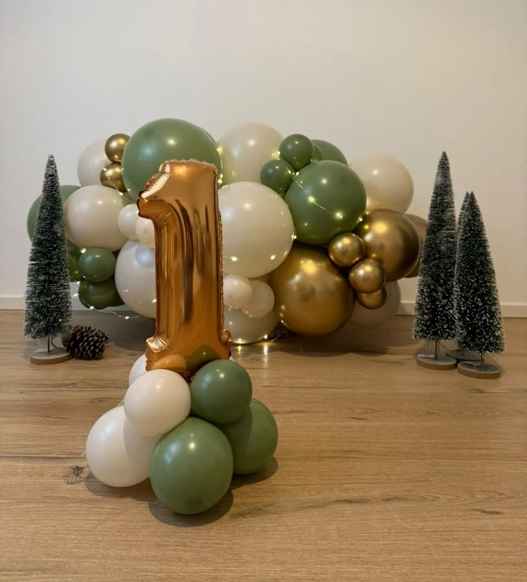 Ghirlanda di palloncini verde salvia, panna e oro con lucine e numero 1 in mylar oro, allestita in casa tra alberelli decorativi