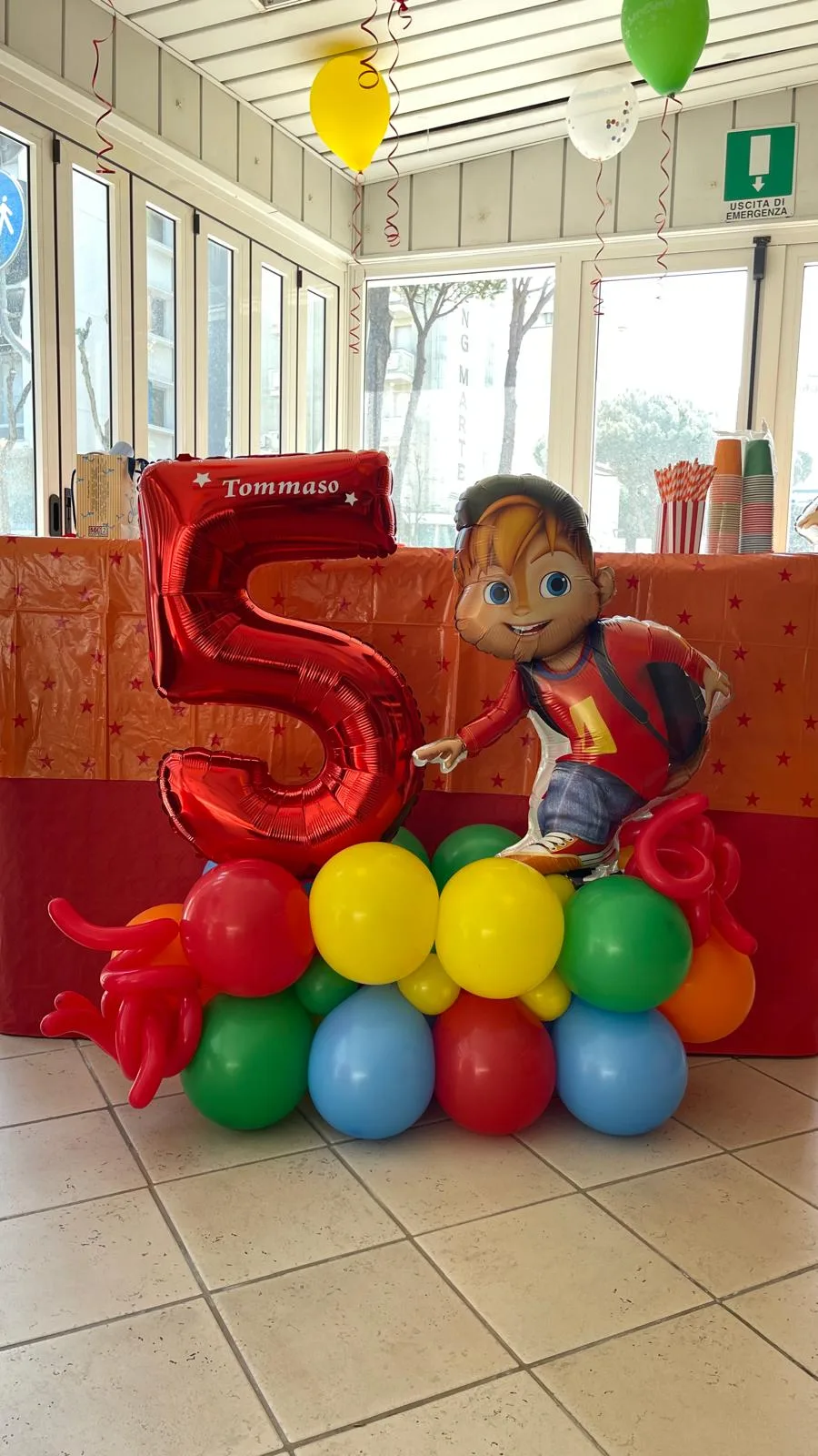 Composizione da terra con numero 5 rosso stampato col nome Tommaso, base di palloncini colorati e sagoma del personaggio dei cartoni