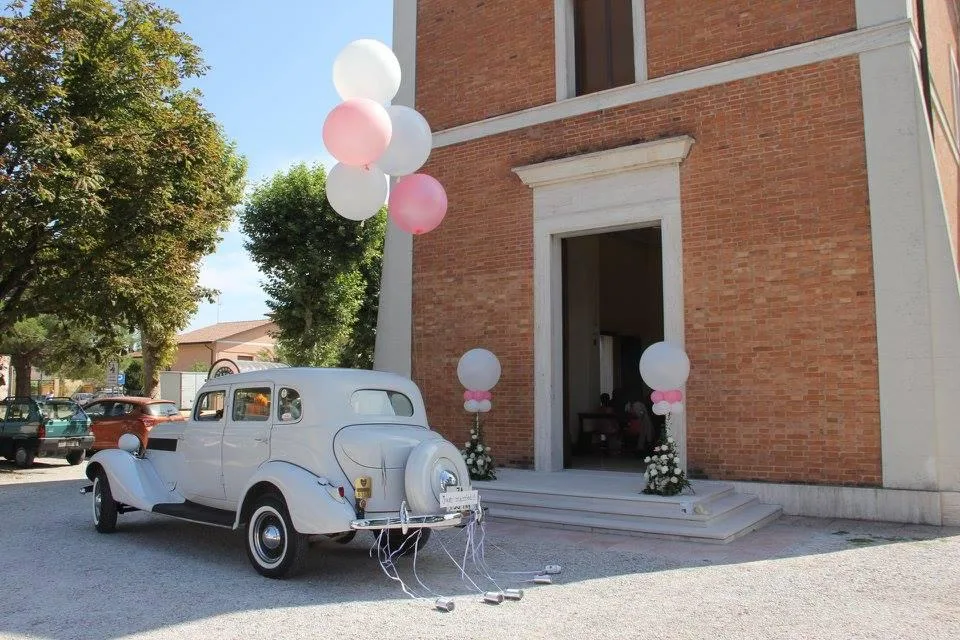 Auto d'epoca bianca degli sposi davanti alla chiesa, con un mazzo di palloncini bianchi e rosa a elio e due colonne di palloncini all'ingresso
