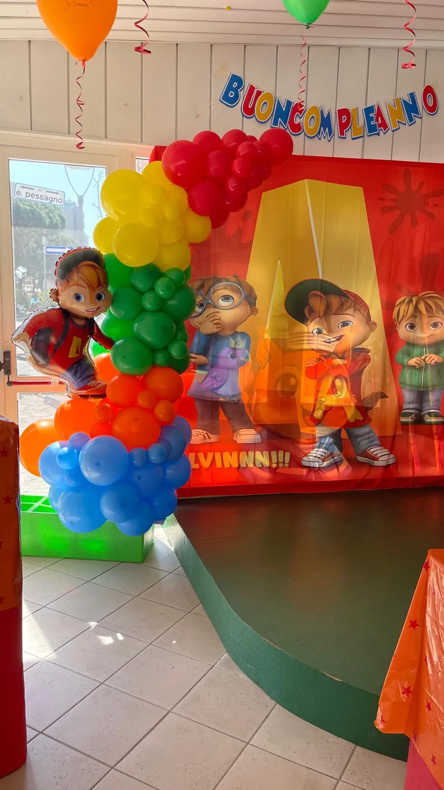Fondale a tema cartoni animati in un locale, con colonna di palloncini arcobaleno, sagoma del personaggio e festone Buon Compleanno