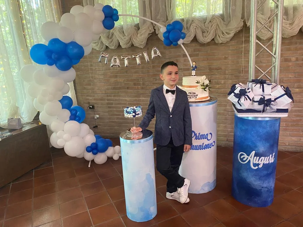 Ragazzo accanto a un arco di palloncini bianchi e blu con il nome Mattia in lettere mylar argento, la torta della Prima Comunione e le colonne stampate con la scritta Auguri