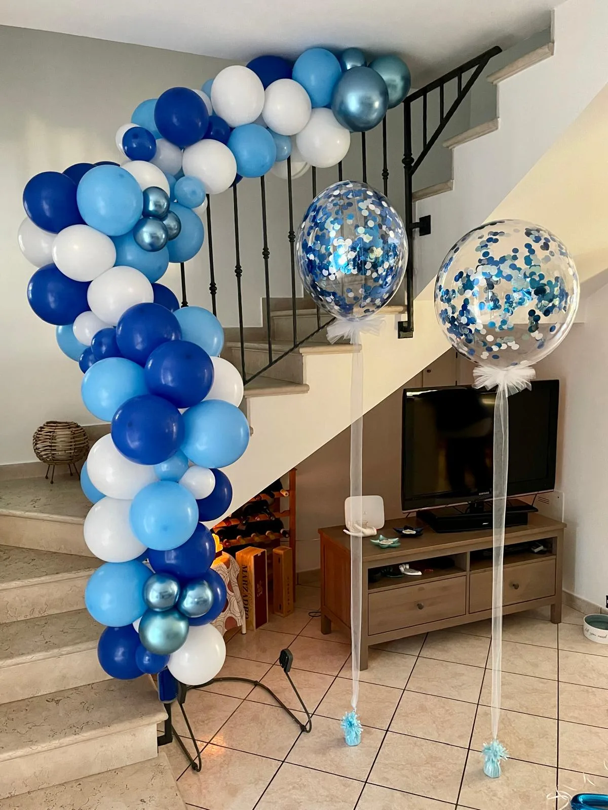 Ghirlanda di palloncini blu, azzurri, bianchi e cromati che sale lungo la scala di casa, con due palloncini trasparenti pieni di coriandoli blu e tulle