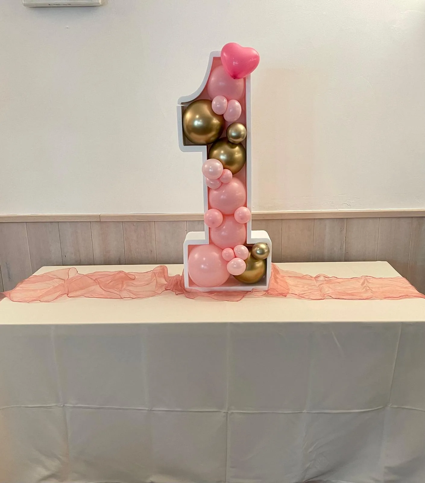 Numero 1 luminoso da tavolo riempito di palloncini rosa e oro con un cuore in cima, su un runner di organza rosa