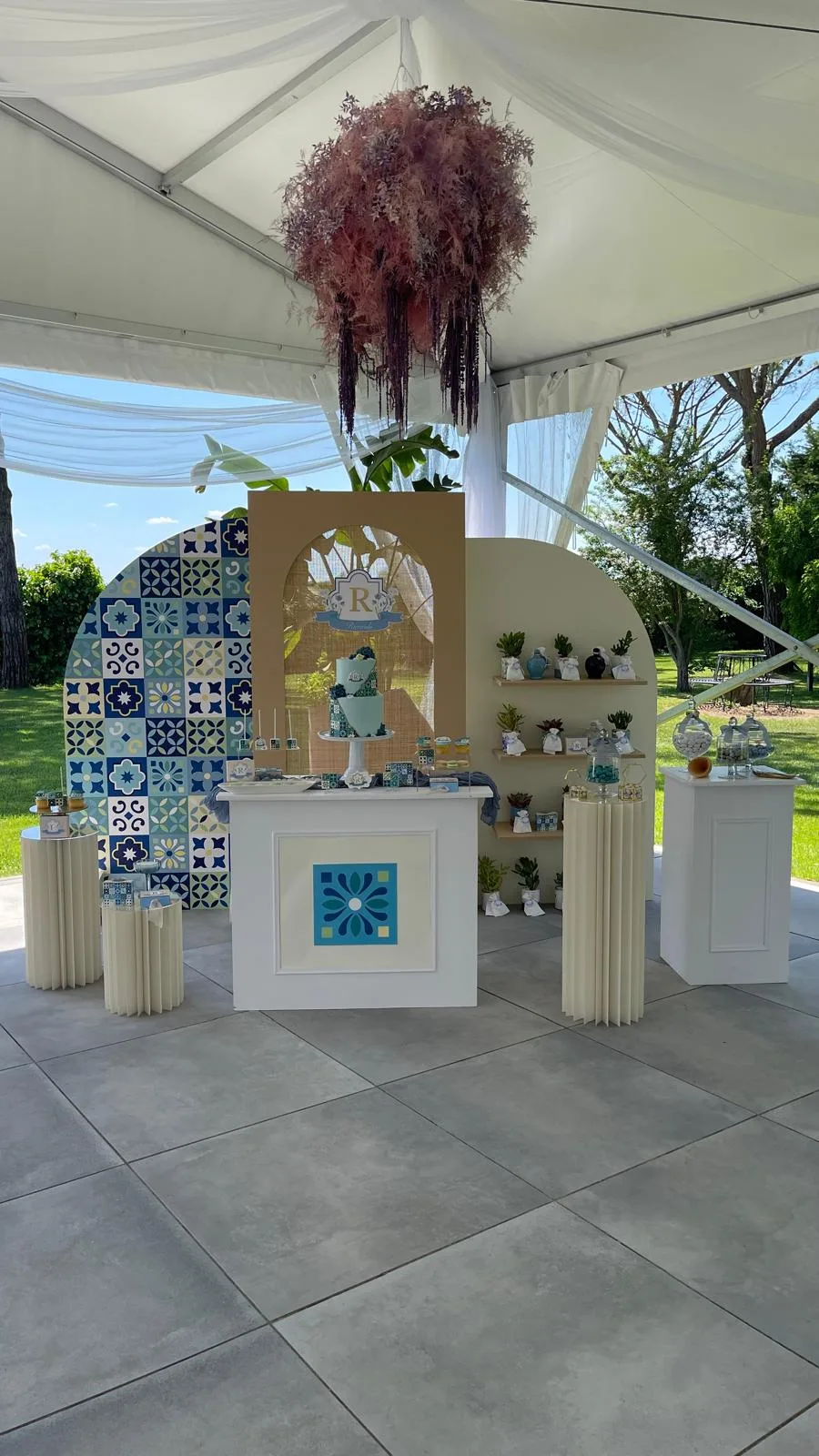 Postazione elegante sotto il gazebo: fondali ad arco con maioliche blu, banco bianco con l'iniziale, mensole dei ricordini, torta e confettata