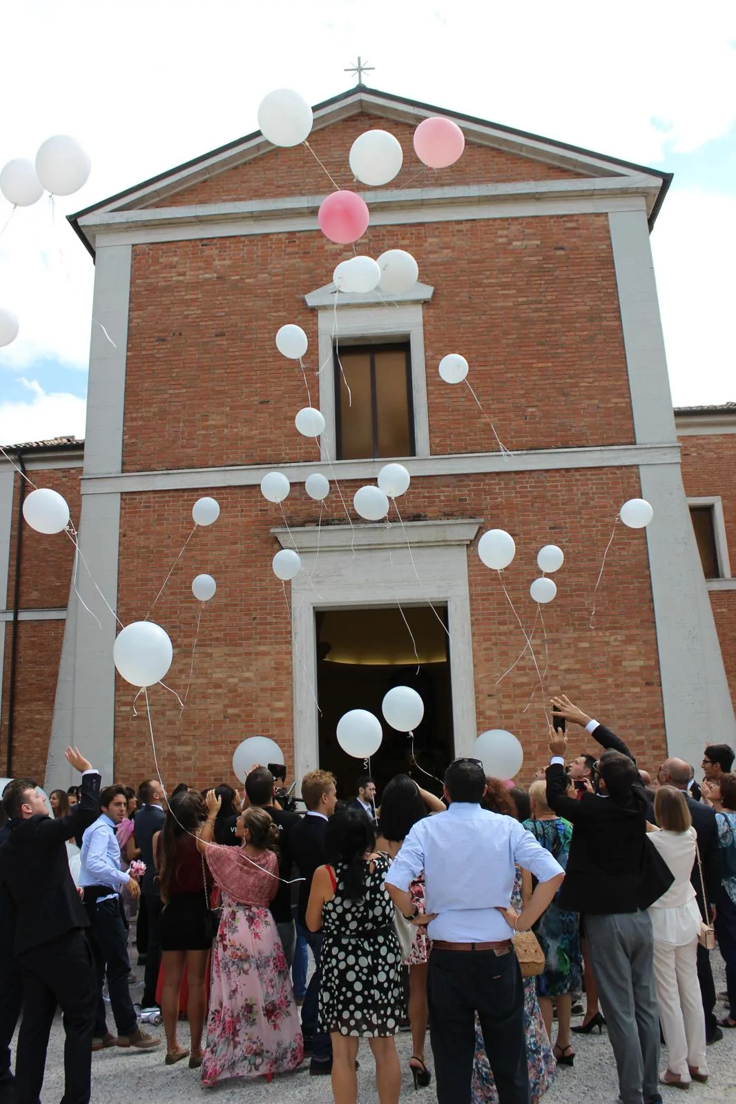 Invitati che lanciano in cielo i palloncini bianchi e rosa davanti alla facciata della chiesa, all'uscita degli sposi