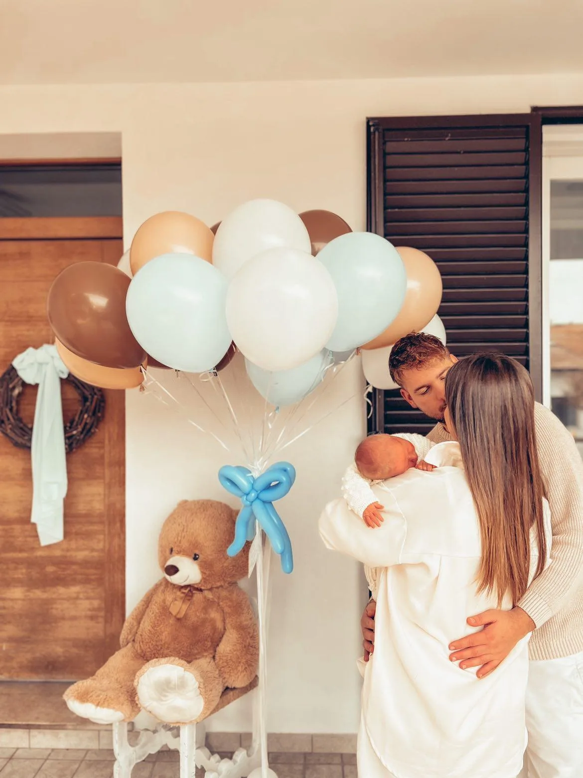 Mamma e papà con il neonato in braccio davanti a casa, accanto a un mazzo di palloncini a elio azzurri, panna e beige con il fiocco fatto di palloncini