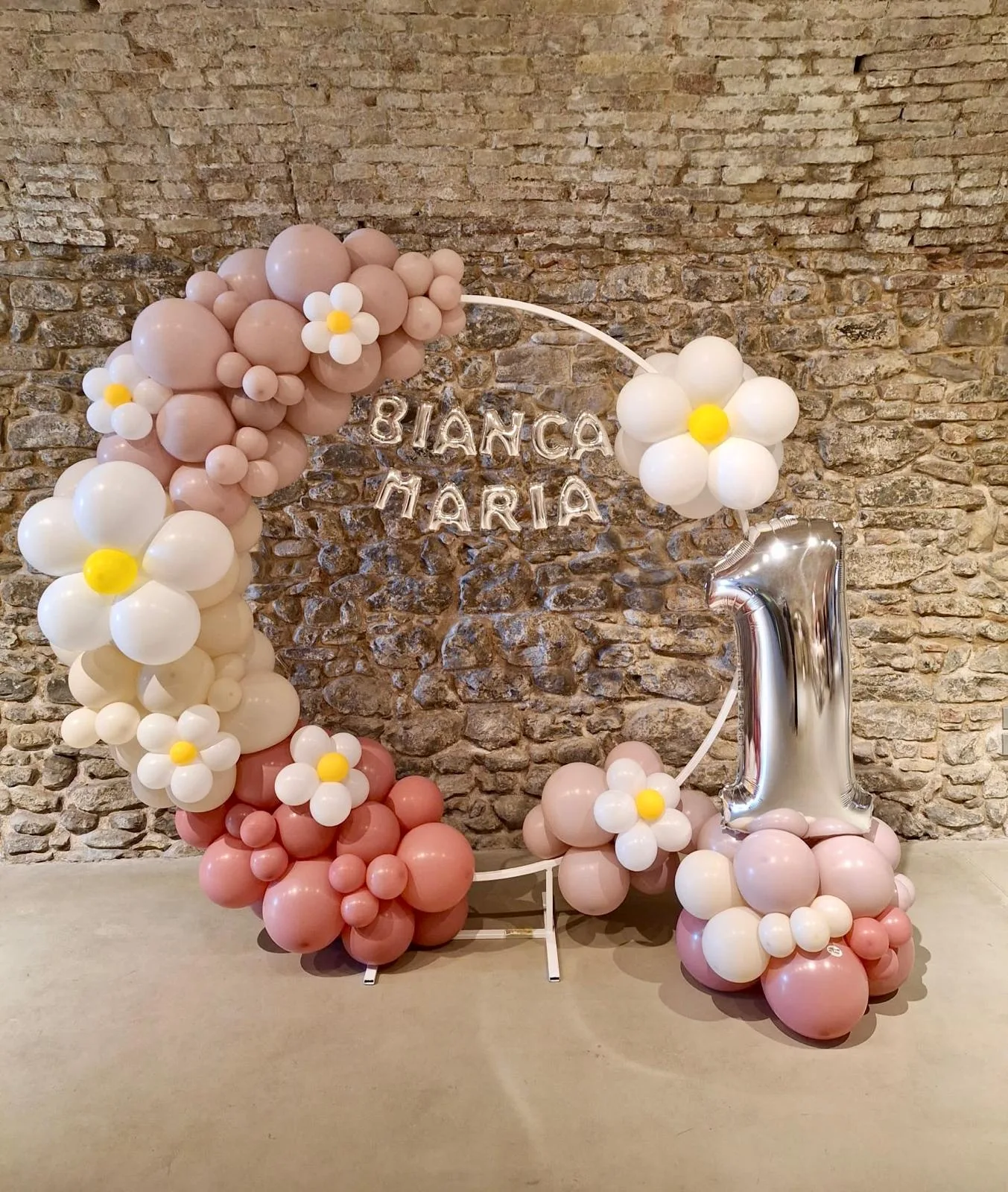 Cerchio di palloncini rosa antico e margherite con le lettere mylar del nome Bianca Maria e numero 1 argento, davanti a un muro in sasso