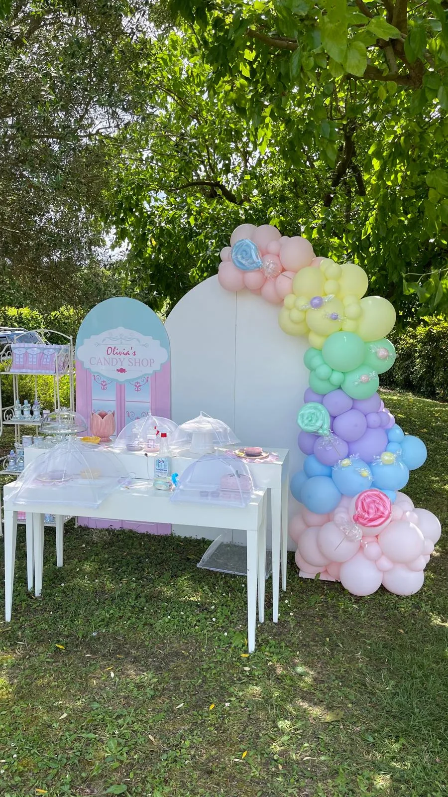 Postazione candy shop in giardino: insegna sagomata col nome Olivia, fondale bianco, ghirlanda di palloncini pastello con caramelle e tavolo dei dolci