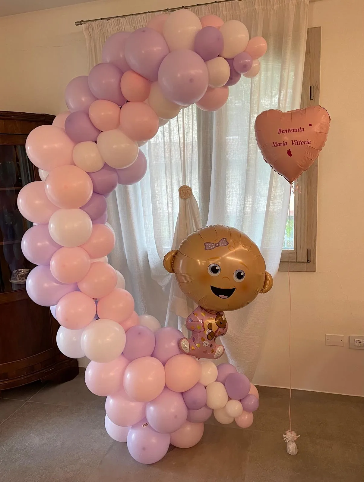 Arco organico di palloncini rosa, lilla e pesca con il palloncino della bimba in mylar al centro e un cuore rosato stampato Benvenuta Maria Vittoria