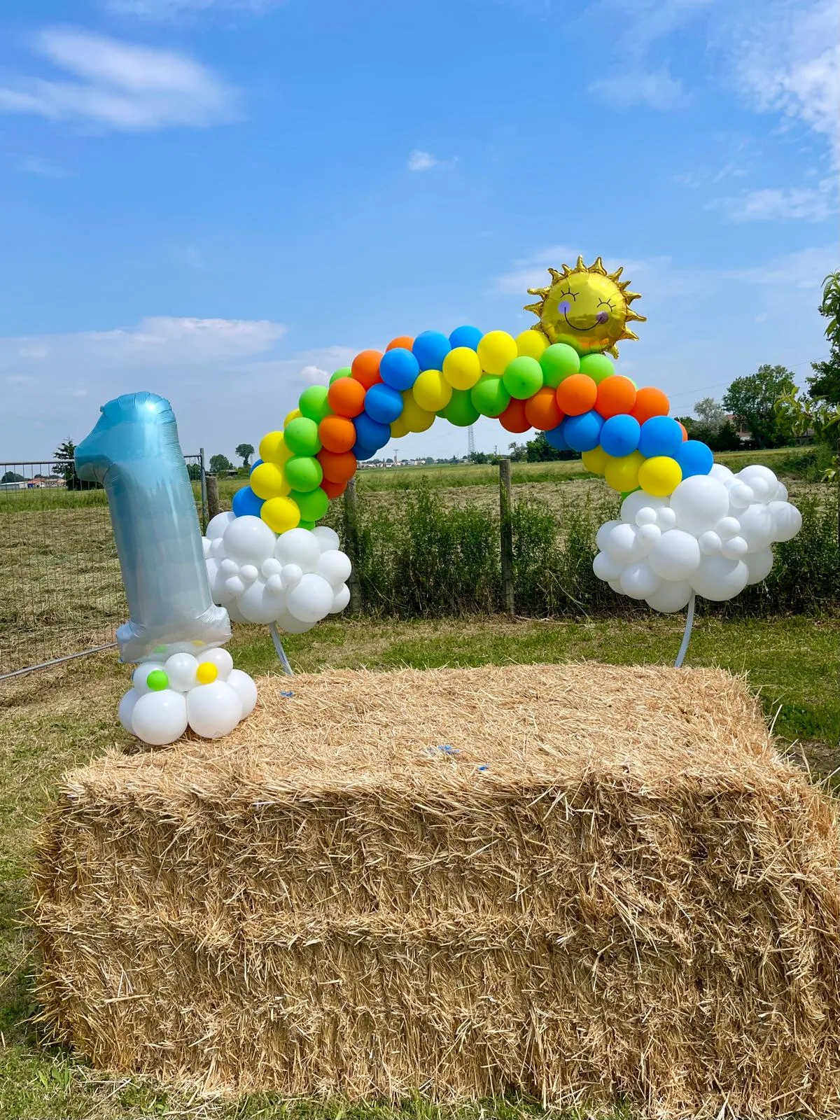 Arco di palloncini arcobaleno con sole e nuvole montato in un prato, con numero 1 celeste accanto a una balla di paglia