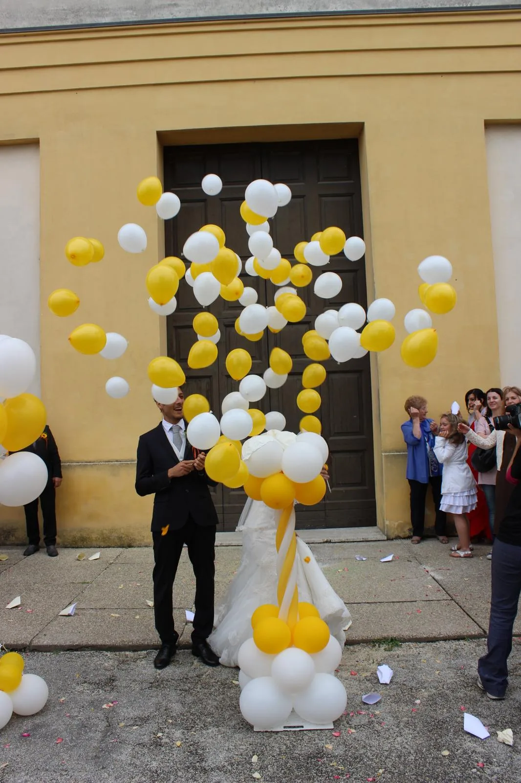 Sorpresa all'uscita dalla chiesa: una sagoma di abito da sposa che libera una nuvola di palloncini bianchi e gialli davanti allo sposo