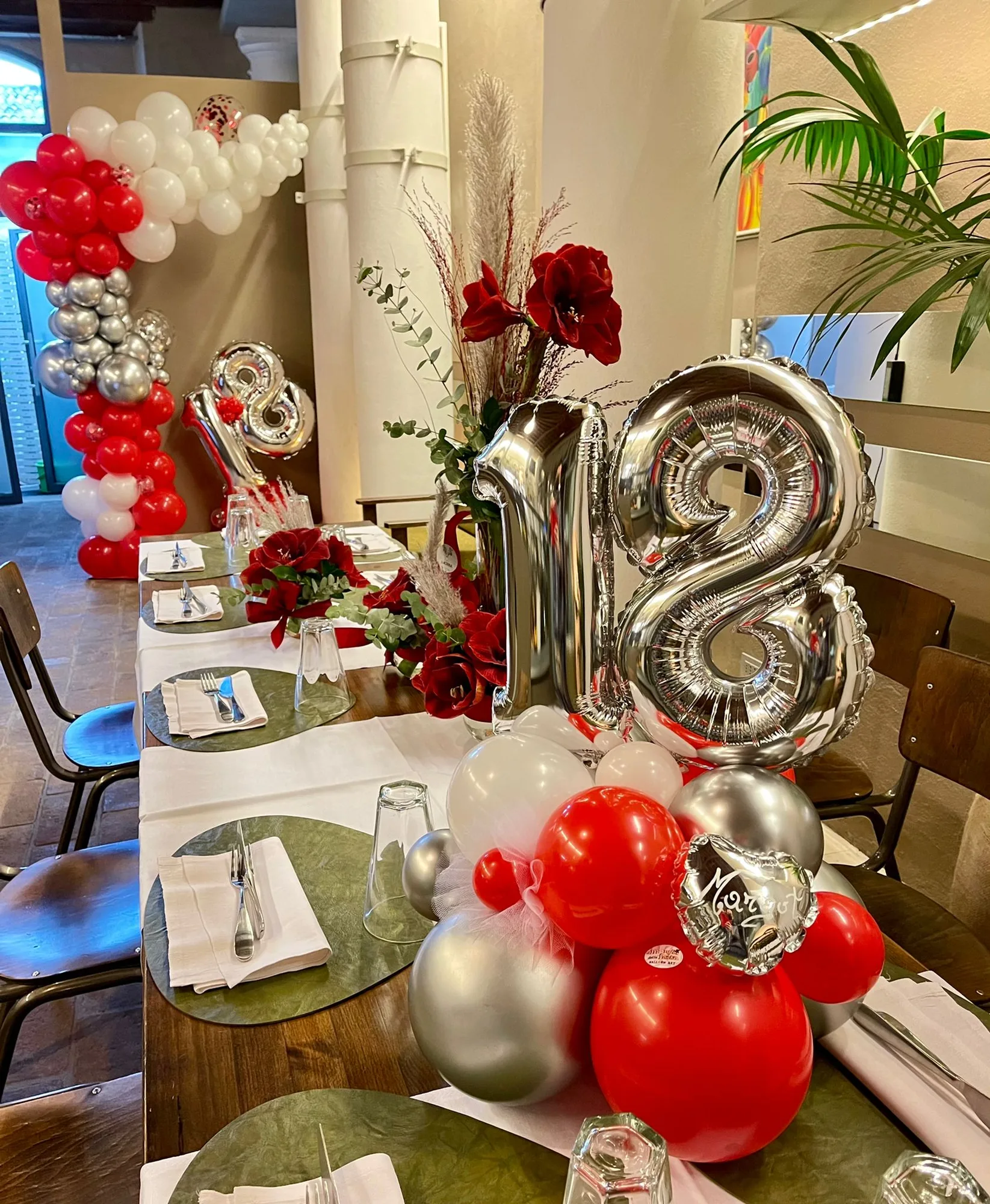 Tavolata di ristorante apparecchiata per un diciottesimo con centrotavola di palloncini rossi, bianchi e argento, numero 18 in mylar e colonna di palloncini all'ingresso