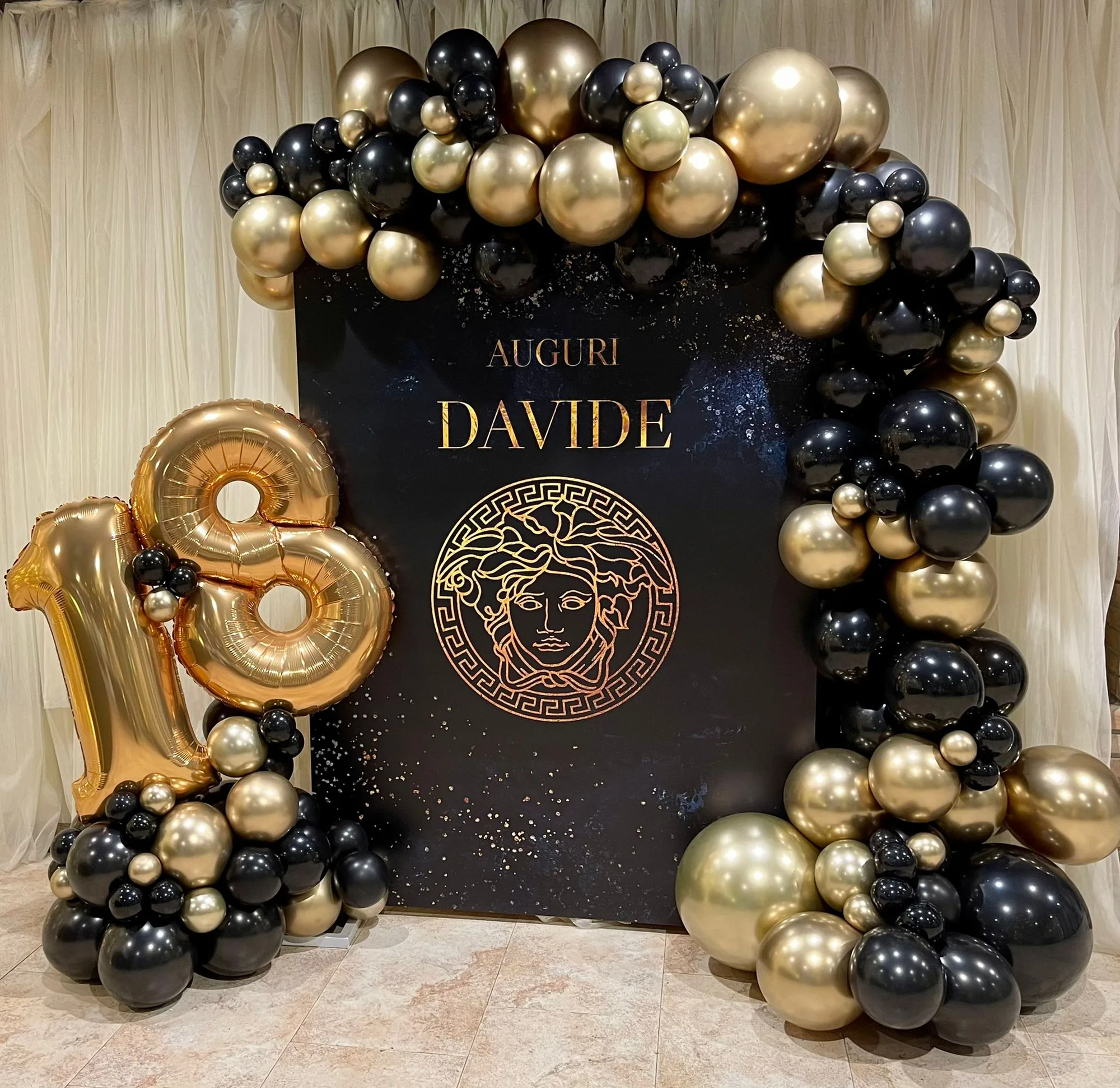 Pannello nero con la scritta dorata Auguri Davide, arco di palloncini neri e oro cromato e numero 18 gigante in mylar oro
