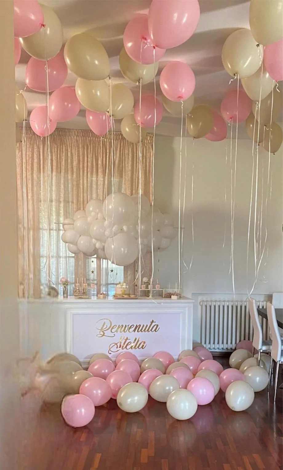 Soffitto coperto di palloncini a elio rosa e panna con nastri lunghi, nuvola di palloncini bianchi sopra il tavolo dei dolci e scritta dorata Benvenuta Stella