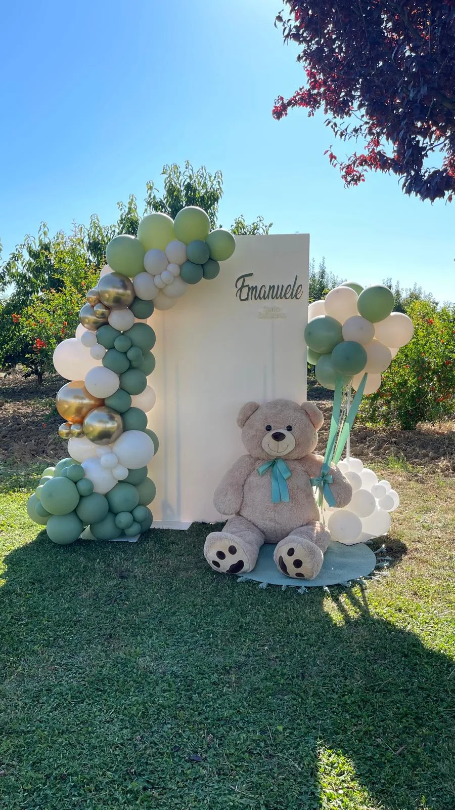 Pannello col nome Emanuele in un frutteto, ghirlanda di palloncini verde salvia, panna e oro e orso di peluche gigante seduto in terra